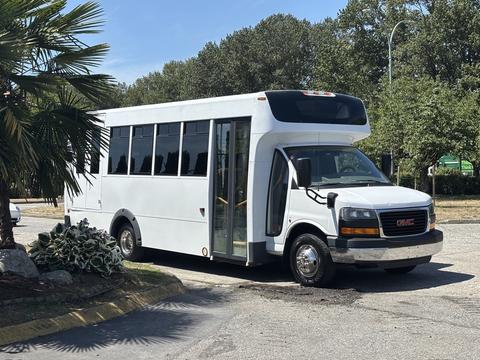 A 2014 GMC Savana bus in white with large windows and a door on the side is parked with its front facing slightly to the right