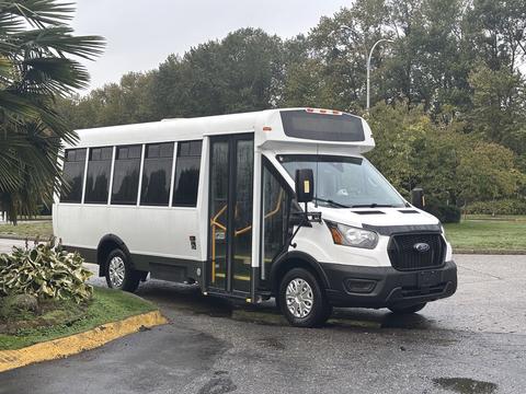 A 2021 Ford Transit-350 bus with a white exterior and large windows has its door open, indicating it's in service for passengers