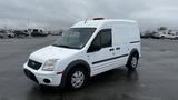 A white 2012 Ford Transit van with a sloped front, sliding side door, and a roof light on top parked in an open area