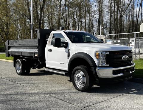 A 2018 Ford F-550 white truck with a flatbed and black dump body parked on a street