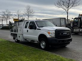 A 2014 Ford F-250 SD in white with a flatbed and tool compartments parked on the side of the road