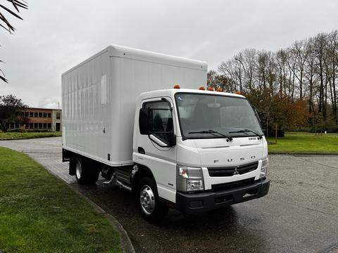 A 2012 Mitsubishi Fuso FE truck with a white box cargo area and an orange light on the roof parked on a paved surface