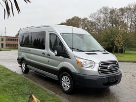 A silver 2019 Ford Transit van with a high roof and large windows parked on a wet surface