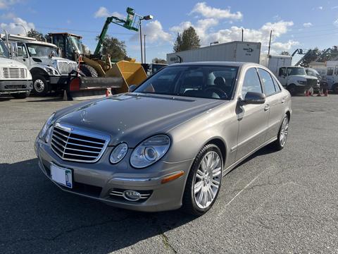 A 2007 Mercedes-Benz E-Class car in a light gold color with chrome wheels parked at an industrial location