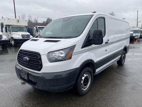 A white 2018 Ford Transit cargo van with a black grille and no side windows parked on a wet surface