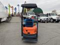 A 2024 AGT L12R forklift with an orange body and blue seat is parked, featuring a protective overhead canopy and a hydraulic lifting mechanism