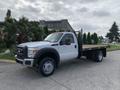 A white 2012 Ford F-550 flatbed truck with a black flatbed and a mesh tailgate parked beside a road