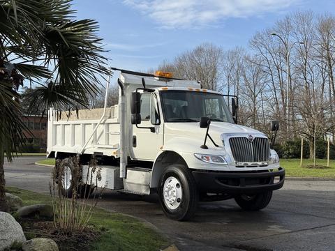 A white 2015 International DuraStar 4300 dump truck with a raised bed and an orange light bar on the roof parked on a street