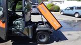 A 2013 Kubota RTV 1100 utility vehicle with an orange dump bed tilted upward and a grey cabin showcasing the driver's seat and dashboard