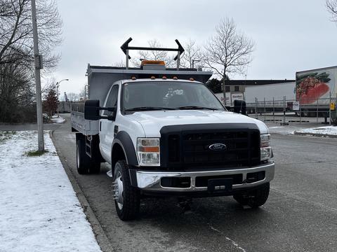 A white 2008 Ford F-450 Super Duty truck with a flatbed and orange warning lights on the roof parked on a snow-covered street