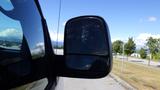 A side mirror of a 2015 GMC Savana reflecting the landscape with a clear view of nearby trees and clouds