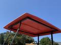 A red canopy structure with a flat roof supported by metal beams viewed from beneath against a clear blue sky