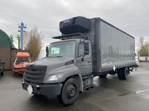 A 2018 Hino 308 box truck with a gray exterior and rear refrigeration unit parked in an industrial area