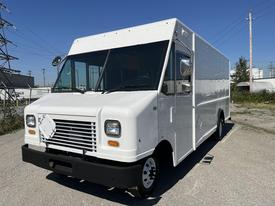 A white 2012 Ford Econoline truck with a boxy shape and large front windows parked on a gravel surface