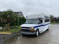 A 2015 Chevrolet Express shuttle bus with a white body and blue stripe parked on a wet road