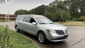 A silver 2013 Lincoln MKT hearse with a soft top parked on a road