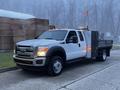 A white 2015 Ford F-550 with a flatbed and toolboxes is parked with its lights on and visible signage on the side