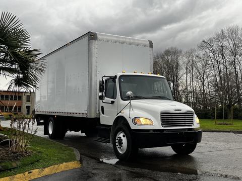 A 2018 Freightliner M2 106 box truck with a white cargo area and a black cab is parked at an angle with its headlights on and several lights shining on the cab roof