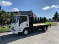 A white 2015 Isuzu NPR truck with a flatbed and a black storage box on the side parked on a street