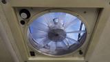 A circular ventilation fan with transparent blades and a mesh cover, mounted in a ceiling unit, showing blue sky through the blades