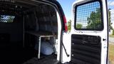 A 2015 GMC Savana cargo van with an open rear door showing a wooden shelf and a metal mesh barrier inside the cargo area