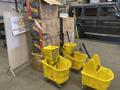 Ten yellow mop buckets and assorted brooms are arranged in the foreground, with some buckets stacked and a caution sign displayed on one of them