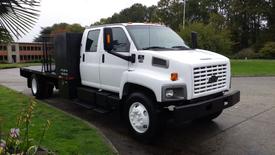 A white 2008 Chevrolet C7500 truck with a black flatbed and tool box on the back has orange clearance lights and features a large front grille with vertical slats