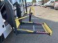 A folding wheelchair ramp in yellow and black extends from the side of a white vehicle with safety stripes and a control button for operation