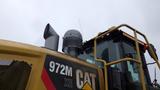 A 2019 Caterpillar 972M XE wheel loader with a prominent exhaust stack and a large glass cab visible in the foreground