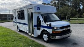 A 2015 Chevrolet Express van designed for accessibility features a white exterior with blue stripes and is equipped with a wheelchair lift on the side