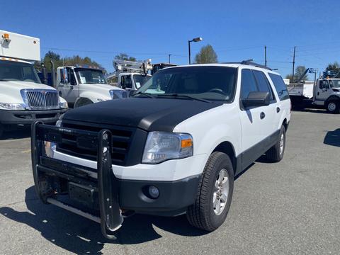 A 2013 Ford Expedition in white with a black hood and front grill guard parked with large trucks in the background