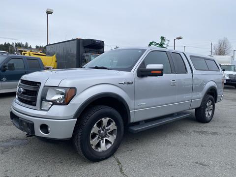 A silver 2014 Ford F-150 with a camper shell is parked beside other vehicles showcasing its prominent front grill and distinctive body style