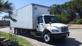 A 2015 Freightliner M2 106 truck with a white box trailer and black front grille parked at a location with greenery visible in the background