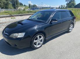 A black 2005 Subaru Legacy Right Hand Drive with a sleek design and five-spoke alloy wheels is parked on a gravel surface