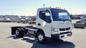 A 2015 Mitsubishi Fuso FE truck with a white body and a flatbed frame positioned in the foreground