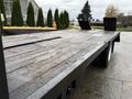 A flatbed trailer with a wooden surface and black steel frame featuring two ramps at the rear for loading and unloading vehicles