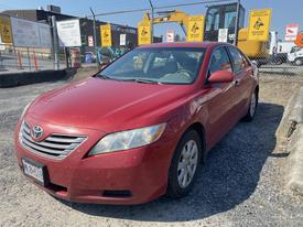 Red 2007 Toyota Camry Hybrid parked in an outdoor area with visible scratches and dirt on the body and a California license plate