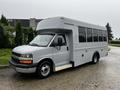 A 2015 Chevrolet Express cutaway bus with a white exterior and large windows on the side is parked on a wet surface with its front facing the viewer