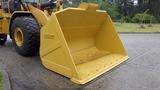 A yellow Caterpillar 972M XE loader bucket positioned on a flat surface with a wide, open front and bolted edge for lifting and carrying materials