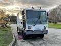 A 2007 Tennant Sentinel street sweeper with a white body and large front windshield parked on a paved surface with its brushes extended for cleaning