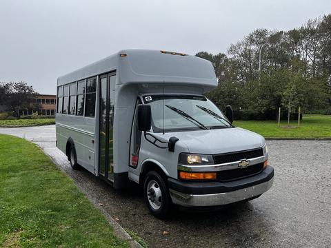 A 2015 Chevrolet Express bus with a gray exterior featuring large windows and a prominent front grille is parked on a wet surface