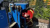 A blue flat deck trailer with visible hydraulic hoses and a red reel system for equipment storage at the rear