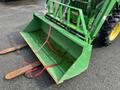 A green 2003 John Deere 790 tractor with a front loader attachment featuring a large bucket secured with red straps and two rusted forks extending from the bottom