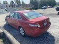 2007 Toyota Camry Hybrid parked on gravel with a red exterior and five-spoke wheels viewed from the rear at an angle