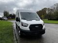 A 2021 Ford Transit van with a white exterior and black grille parked on a street with greenery nearby