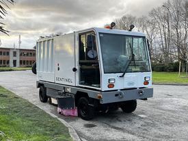 A 2007 Tennant Sentinel Vacuum with a white body and large front windows designed for street cleaning featuring a side broom and safety lights on top