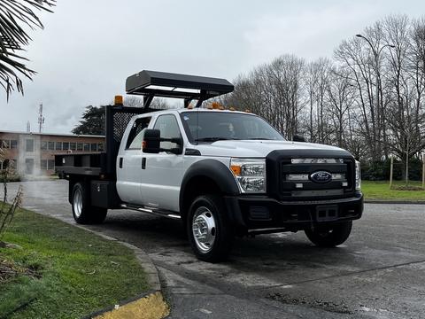 A white 2015 Ford F-550 flatbed truck with a tool box mounted on the back and a black grille parked on a road