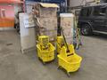 Ten yellow mop buckets and assorted brooms are arranged in the foreground with several cardboard boxes in the background