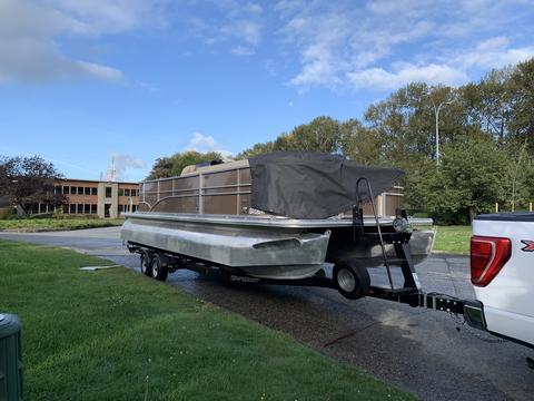 A 2013 Sylvan 13 person boat on a trailer with a cover over the seating area near a road and grassy area