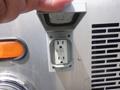 A hand is opening a gray electrical outlet cover revealing a three-prong outlet on a silver vehicle surface